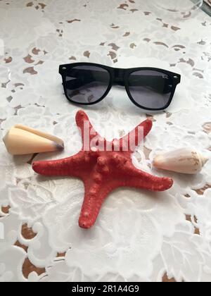 Ein wunderschöner roter Seestern und eine schwarze Sonnenbrille liegen mit Muscheln auf weißem Stoffhintergrund dekoriert. Tropischer Sommer mit Meeresumwelt. Stockfoto