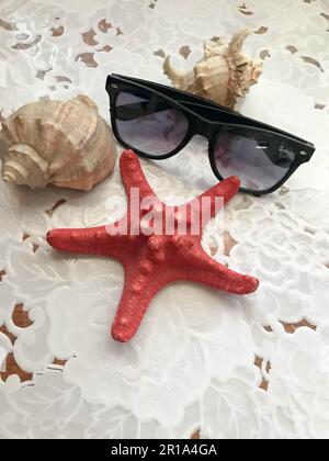 Ein wunderschöner roter Seestern und eine schwarze Sonnenbrille liegen mit Muscheln auf weißem Stoffhintergrund dekoriert. Tropischer Sommer mit Meeresumwelt. Stockfoto