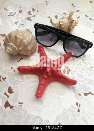 Ein wunderschöner roter Seestern und eine schwarze Sonnenbrille liegen mit Muscheln auf weißem Stoffhintergrund dekoriert. Tropischer Sommer mit Meeresumwelt. Stockfoto