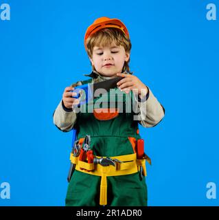 Kind im Schutzhelm und Werkzeuggürtel mit Säge. Mit Werkzeugen arbeiten. Ein Junge in Bauernuniform mit Werkzeug für die Reparatur. Kleiner Handwerker oder so Stockfoto