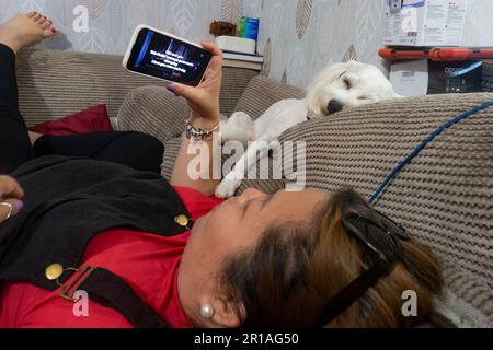 Eine Dame entspannt sich abends auf einem Sofa und sieht sich Videos auf ihrem Handy an, während ihr kleiner weißer Cavapoo neben ihr schläft. Stockfoto