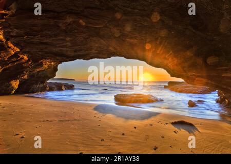 Helle aufgehende Sonne über dem Horizont des Pazifischen Ozeans aus dem Inneren der Meereshöhle in Caves Beach in Australien. Stockfoto