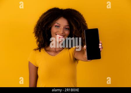 afro-Frau mit lockigen Haaren, die auf das Handy auf gelbem Hintergrund zeigen Stockfoto