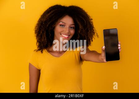 afro-Frau mit lockigen Haaren, die auf das Handy auf gelbem Hintergrund zeigen Stockfoto