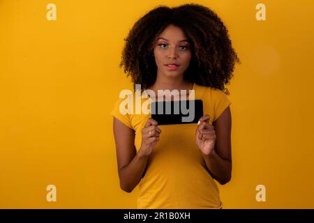 afro-Frau mit lockigen Haaren, die auf das Handy auf gelbem Hintergrund zeigen Stockfoto