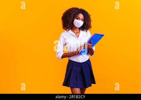 Afro-Mädchen mit Büchern auf gelbem Hintergrund und Schutzmaske. Kehren Sie in Quarantäne zu den Klassen zurück Stockfoto
