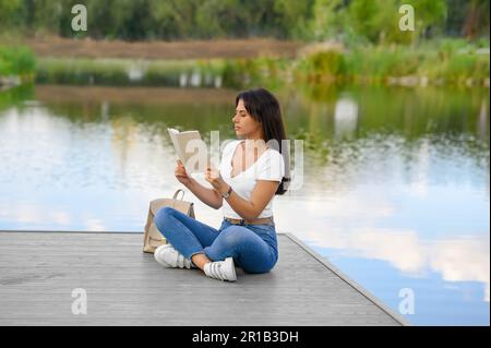 Junge Frau liest Buch auf dem Pier in der Nähe des Sees Stockfoto