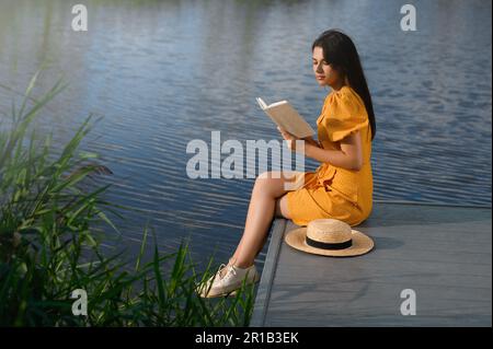 Junge Frau liest an sonnigen Tagen ein Buch in der Nähe des Sees Stockfoto