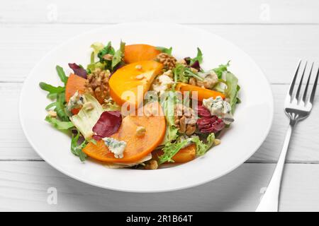 Köstlicher Persimonsalat und Gabel auf weißem Holztisch, Nahaufnahme Stockfoto