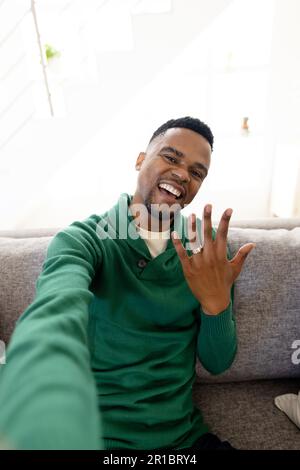 Porträt eines glücklichen afroamerikanischen Schwulen, der Ehering zeigt, während er im Wohnzimmer auf dem Sofa sitzt Stockfoto