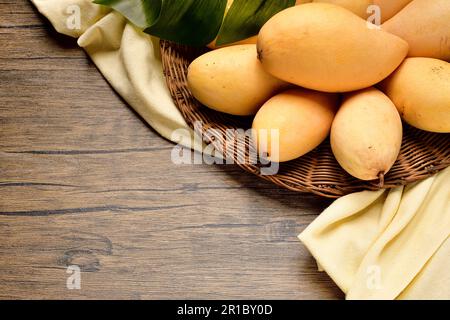 Blick von oben viele Mangos in Korb mit Korb aus Korb auf Holztisch. Thailändische Mangos und Stoffe auf Holzhintergrund. Gelbe Mangos von oben auf Holztisch Stockfoto