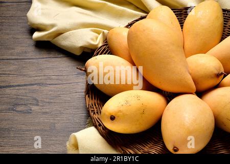 Blick von oben viele Mangos in Korb mit Korb aus Korb auf Holztisch. Thailändische Mangos und Stoffe auf Holzhintergrund. Gelbe Mangos von oben auf Holztisch Stockfoto