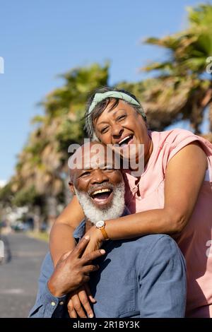 Portrait eines glücklichen älteren afroamerikanischen Ehepaars, das sich auf der Promenade über Palmen umarmt Stockfoto