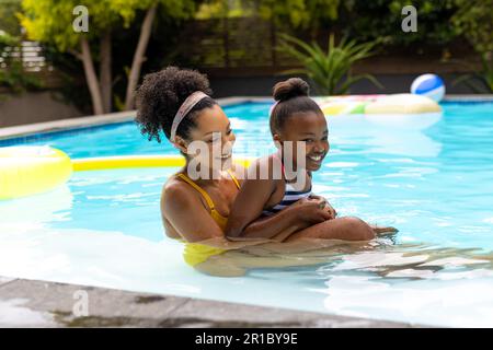 Glückliche afroamerikanische Mutter und Tochter, die Spaß zusammen haben, sich im Swimmingpool zu umarmen Stockfoto