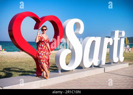 Rothaarige Frau mit Sonnenbrille, die sich auf ein großes rotes 3D-Herz lehnt und in der Nähe des „Sarti“-Textes platziert ist. Willkommen im Sarti-Dorf in Griechenland. Stockfoto