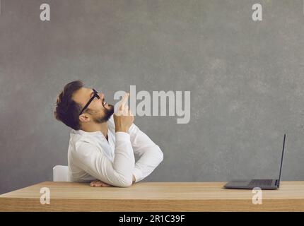 Ein junger Mann mit Bart und Brille sitzt mit einem Laptop an einem Tisch und zeigt seinen Zeigefinger nach oben Stockfoto