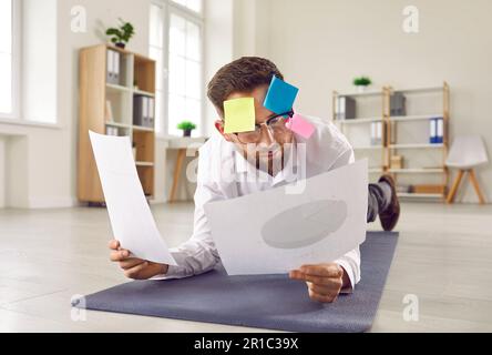 Lustiger Mann mit Haftnotizen, Multitasking im Gesicht, Arbeiten mit Papieren und Übungen auf Yogamatte Stockfoto