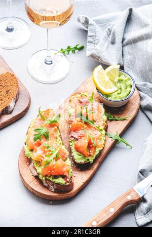 Gourmet-Mittagessen mit Avocado-Lachs-Toasts auf Roggenbrot und Rosenweingläsern, grauer Tischhintergrund, Blick von oben Stockfoto
