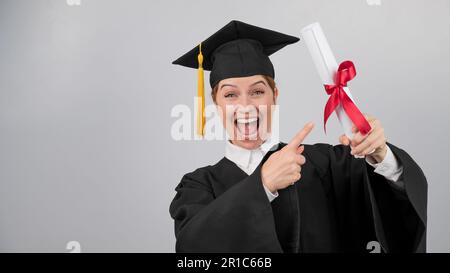 Glückliche Frau im Graduiertenkleid, die mit dem Finger auf ein Diplom zeigt. Stockfoto