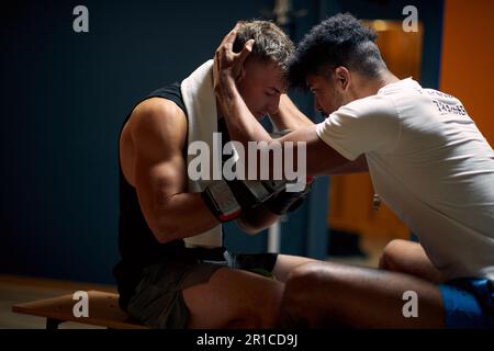 Der Trainer hilft dem jungen Boxspieler dabei, sich mental vorzubereiten, seinen Kopf zu halten und mit ihm zu sprechen, in der Garderobe, die auf einer Bank sitzt. Sport, Kampfsport, Leben Stockfoto