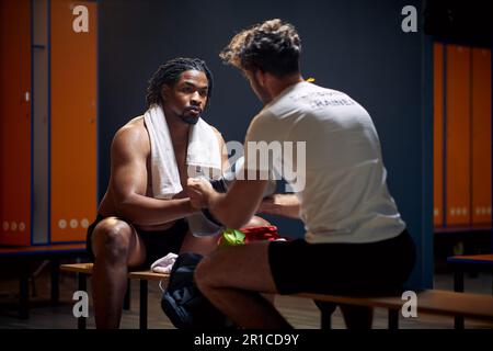 Starker junger Boxspieler nach dem Workout mit dem Trainer, der ihm dabei hilft, die Handschuhe auszuziehen, und der sich im Gespräch von Angesicht zu Angesicht in der Garderobe aufhält. Sport, Lifestyle Stockfoto