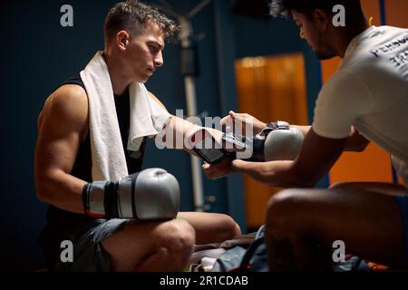 Junger Boxspieler, der sich auf das Boxtraining vorbereitet, der Trainer hilft ihm beim Anziehen von Boxhandschuhen. Sport, Lifestyle, Kampfsport-Konzept. Stockfoto