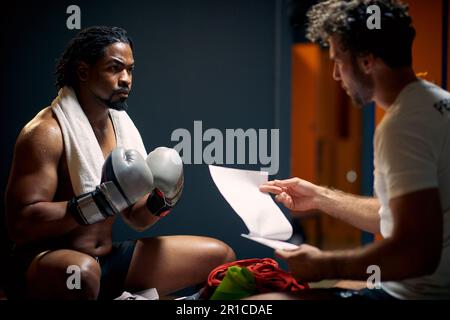 Muskulöser und starker Boxspieler in Schweiß nach dem Workout, der sich von Angesicht zu Angesicht auf der Bank mit seinem Trainer ruht, um Beratung zu erhalten. Sport, Lifestyle-Konz Stockfoto