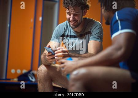 Junger Mann mit Handy und Freund, der auf der Bank im Umkleideraum des Fitnessstudios sitzt und vor dem Training Spaß hat. Sport, Gesundheit, Zusammengehörigkeitskonzept. Stockfoto