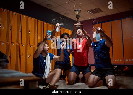Eine Gruppe von Basketballspielern hält die goldene Trophäe hoch und feiert den Erfolg in der Garderobe, schreit und spritzt Wasser. Erster Platz gewonnen. Stockfoto