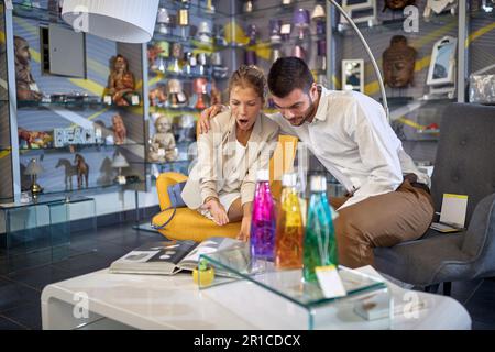 Ein junges Paar sitzt zusammen in einem Lampengeschäft und durchsucht einen Katalog von Armaturen. Die Frau hat eine Seite umgedreht und zeigt aufgeregt auf mich Stockfoto