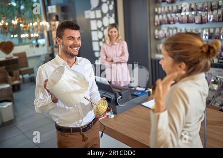 Ein Paar in einem Laden, das eine beeindruckende Tischlampe bewundert. Der Mann zeigt es seinem Partner, der beeindruckt und glücklich aussieht. Eine lächelnde Verkäuferin sieht mich Stockfoto