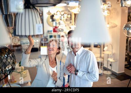 Ein junges Paar in einem Beleuchtungsgeschäft, das mit Begeisterung und Freude im Gesicht durch Kronleuchter und Lampen schlendert. Stockfoto