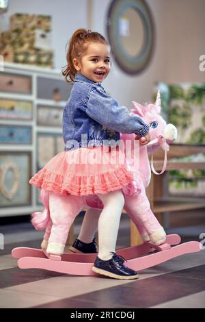 Glückliches und fröhliches kleines Mädchen, das auf einem schaukelnden Einhorn sitzt. Stockfoto