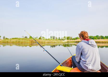 Junge, der im Fischerboot auf dem ruhigen See sitzt und eine Rod hält, in einem malerischen Rückblick auf das friedliche Outdoor-Abenteuer, das eine Frühlingslandschaft schafft Stockfoto