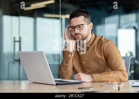 Pensiver und seriöser junger Student, der an einem Schreibtisch in einer Campus-Bibliothek sitzt und online auf einem Laptop lernt. Absolviert eine Prüfung, schreibt einen Test, besteht ein Vorstellungsgespräch, hört sich einen Kurs bei Videogesprächen an. Stockfoto
