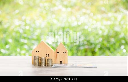 Stapeln Sie Geldmünzen und Dollars auf einem Holztisch mit einem simulierten Holzhaus auf dem im Garten verschwommenen Gras mit einem Kopierraum auf der oberen Seite. Stockfoto