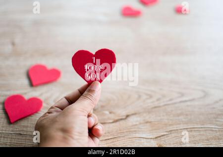 Konzept Valentinstag, Männerhand mit rotem Herzsymbol, Buchstaben Ich liebe dich weiß und rotes Herzsymbol verschwommen auf einem Holztisch. Nahaufnahme Stockfoto