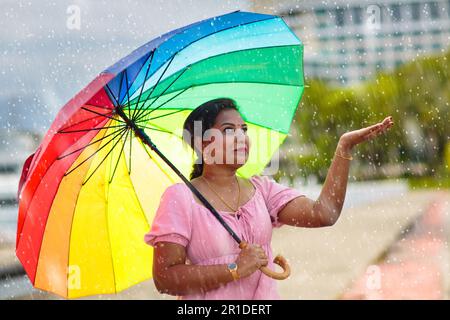 Junge Frau, die unter starkem Regen mit Regenbogenschirm läuft. Eine Indianerin, die sich in der Herbstdusche amüsiert. Herbstferien. Hübsches Mädchen in rosa Kleid Stockfoto