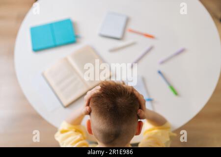 Aus einem großen Winkel von hinten betrachtet ein erschöpfter Schüler, der müde ist, weil er den Kopf gehalten hat und Hände am Schreibtisch sitzen, mit Papier aus dem Copybook, mit Blick nach unten. Stockfoto