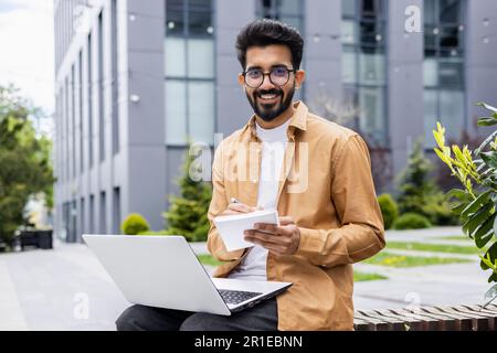 Porträt eines jungen, glücklichen Hindu-Mannes, eines Schülers außerhalb des Campus, der lächelt und in die Kamera schaut, eines Lehrers, der ein Notebook für Online-Lernen verwendet, und Videoanrufe mit Schülern Stockfoto