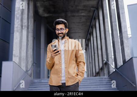 Erfolgreicher junger Hindu-Mann, der vor dem Bürogebäude spaziert, Programmierer für Softwareentwickler, der lächelt und mit der Test-App am Telefon zufrieden ist, zufrieden mit dem Ergebnis. Stockfoto