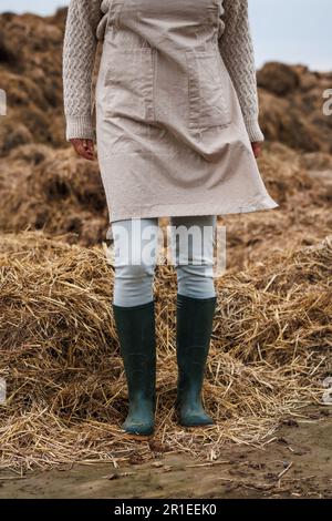Bauernfrau mit Schürze und Gummistiefeln steht neben dem organischen Düngerhaufen. Landwirtschaftliche Tätigkeit im Herbst Stockfoto