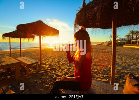 Glückliche asiatische Frau, die Rotwein trinkt und den Sonnenuntergang am Strand beobachtet, Festungskonzept Stockfoto