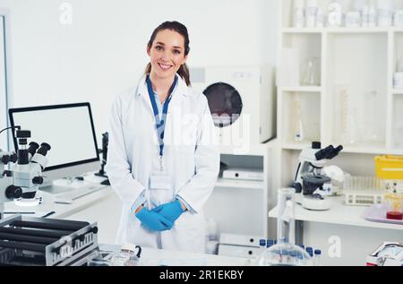 Willkommen in meinem Büro. Porträt einer fröhlichen jungen Wissenschaftlerin, die an ihrem Arbeitsplatz steht und bereit ist, in einem Labor zu arbeiten. Stockfoto