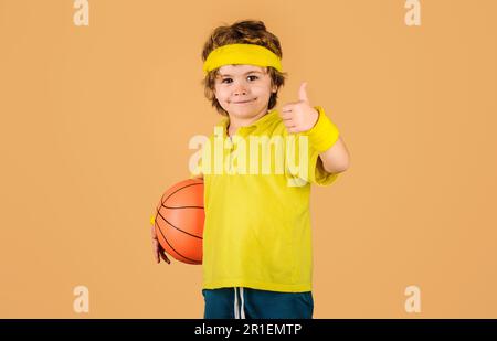 Kleiner Basketballer mit Basketballball, der Daumen nach oben zeigt. Kind in Sportbekleidung mit Basketball. Sportspiel. Aktiver Sport-Lifestyle. Basketball Stockfoto