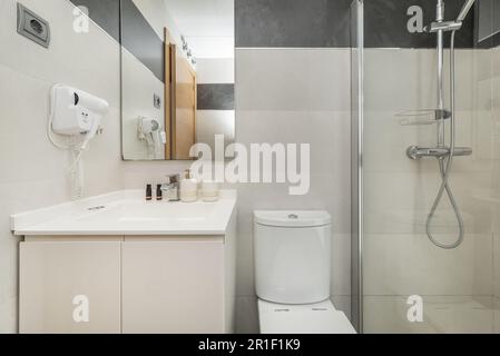 Kleines Badezimmer mit bodenebener Dusche mit Steinboden, weißer Schrank mit Waschbecken, wandmontiertem Haartrockner und Spiegel mit Ablage Stockfoto