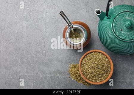 Calabash mit Bombilla, Teekanne und Schüssel Teeblätter auf grauem Tisch, flach liegend. Platz für Text Stockfoto