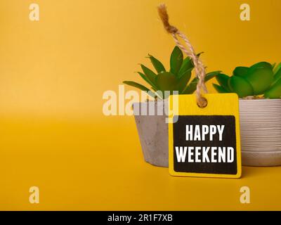 Holzbrett und grüne Pflanze mit dem Wort HAPPY WEEKEND auf gelbem Hintergrund Stockfoto