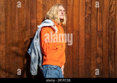 Der entspannte, blonde Mann in der Jacke über der Schulter mit geschlossenen Augen steht in der Sonne an der Holzwand Stockfoto