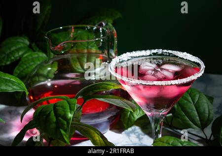Rotes Getränk mit Eis in einem margarita-Glas mit einem Zuckerrand. Im Hintergrund befindet sich eine Kanne mit einem Cocktail, umgeben von grünen Blättern einer Pflanze. Bei einer mutter Stockfoto